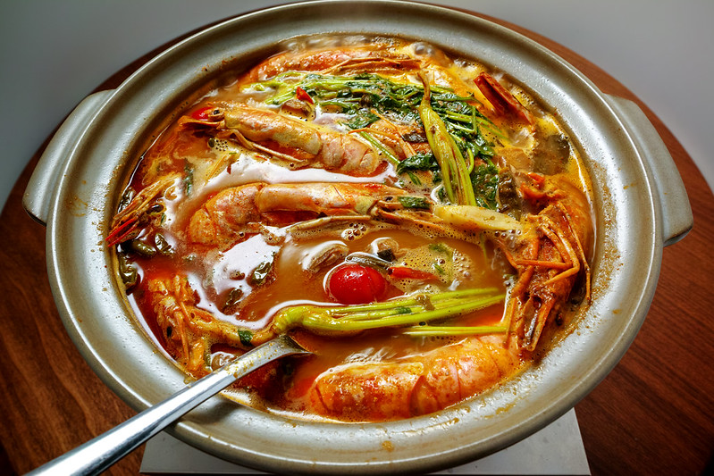 Closeup shot of a hot Tom Yum in a clay pot with a spoon in, prawns with their head and shell still intact, veggies like wilted spinach and tomato.