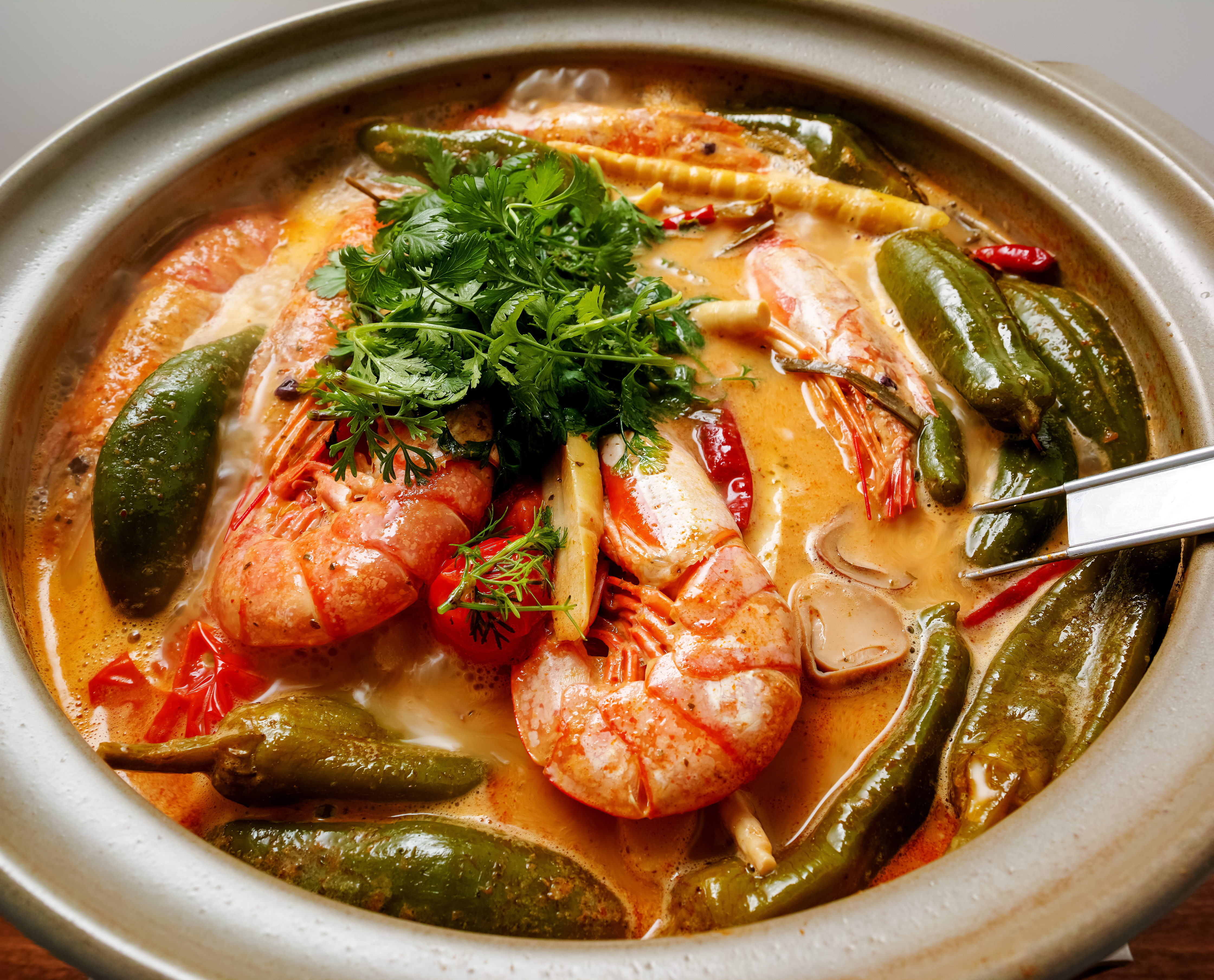 Closeup shot of Tom Yum with chilis and shrimp with peels still on topped with cilantro in a neutral coloured bowl with a metal spoon