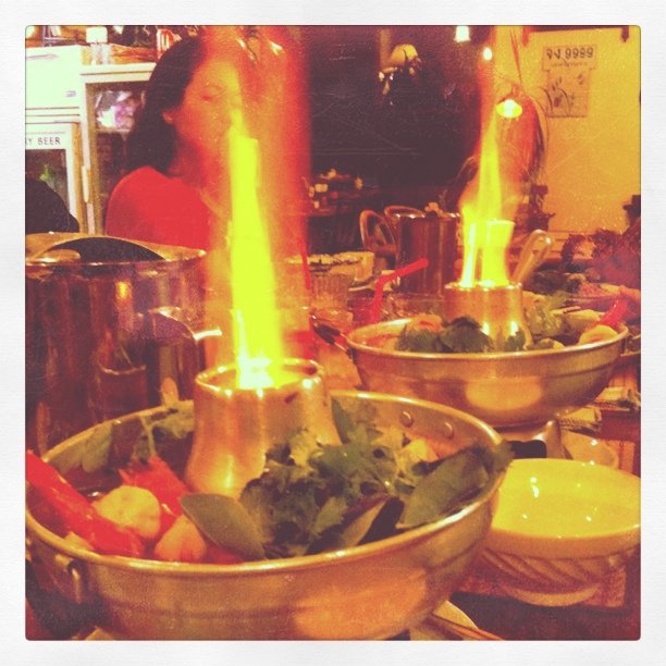 An old photograph at a Thai restaurant with a woman in the back, two fire pots with flares and Tom Yum ingredients in the fire pot bowl, also has bowls and pots seemingly for a hotpot