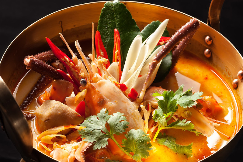 An extreme closeup shot of Tom Yum with mixed seafood inside of a pot topped with a single prawn head, small crab legs, sliced thai chilies, lime kaffir leaves, green onions resembling the leaves and petals of a flower placed intricately around the prawn head, with fish balls and pretty leaves of cilantro on the soup.